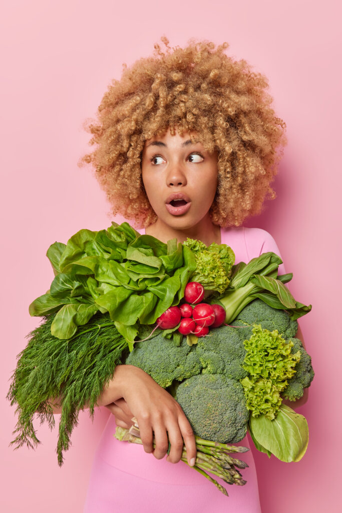 Impressed young curly haired woman stares away shocked. She carries variety of green vegetables picked up directly from field harvest on a farm isolated over pink background. Spring harvest concept